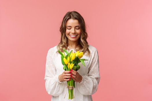 Holidays, beauty and spring concept. Portrait of lovely, romantic blonde girl in white dress, holding yellow tulips, looking at beautiful flowers, smiling and blushing, pink background.