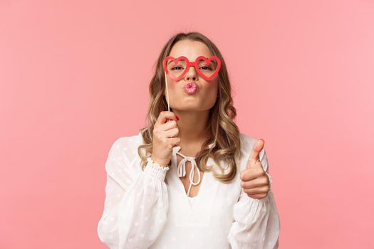 Spring, happiness and celebration concept. Close-up portrait sassy and coquettish attractive blond girl in white dress, holding heart-shaped glasses on eyes, folding lips in kiss and show thumbs-up.