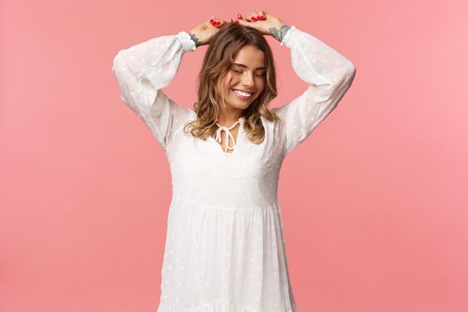 Beauty, tenderness and fashion concept. Attractive blond caucasian woman with tattoos in light white spring dress, raise hands up relaxed smiling with closed eyes, dancing, pink background.