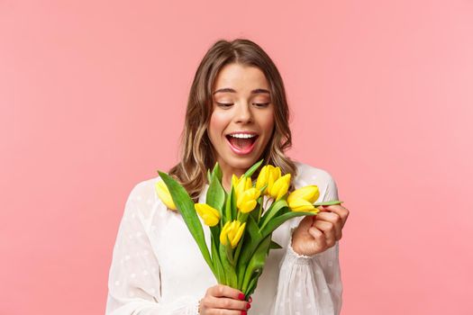 Spring, happiness and celebration concept. Portrait of surprised and pleased happy girl cheerfully looking at bouquet of flowers, smiling amused, receive yellow tulips, stand pink background.