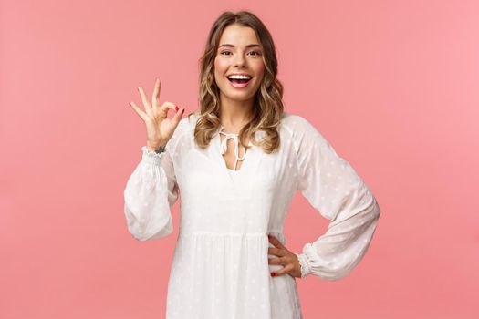 Portrait of carefree good-looking blond girl, wear white dress, guarantee you will like this special spring offer, show okay sign, agree or accept, smiling and nod in approval, pink background.