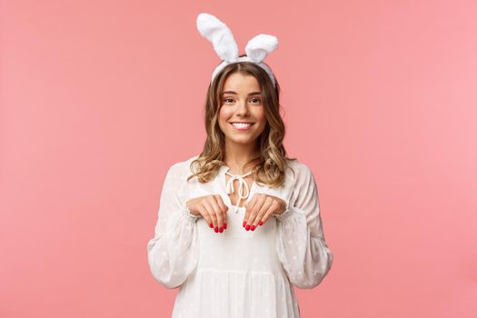 Holidays, spring and party concept. Portrait of cute and tender, lovely blond girl imitating bunny, holding hands like paws and wearing rabbit ears, smiling camera, pink background.