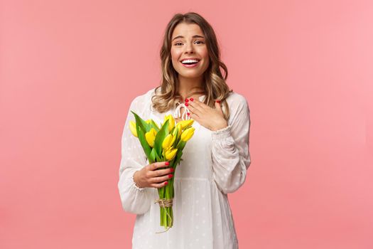 Holidays, beauty and spring concept. Grateful charming blond girl receive yellow tulips, feel touched thanking for romantic gift, sighing lovely smiling, feel happy hold flowers, pink background.