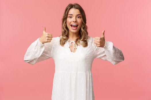 Portrait of feminine blond woman in white dress, show thumbs-up in approval, say yes, approve or agree, satisfied and recommend product, standing pink background in white dress.