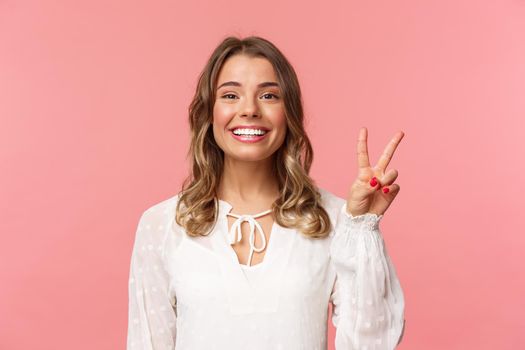 Close-up portrait of romantic lovely smiling girl with blond short hair, wearing white dress, show peace sign enjoying spring, grinning and look camera over pink background. Copy space