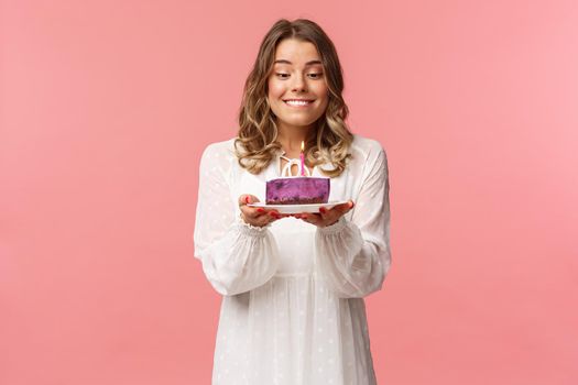 Holidays, spring and party concept. Portrait of dreamy, happy birthday girl feeling excitement and joy celebrating b-day, biting lip and smiling as making wish blowing candle on cake.