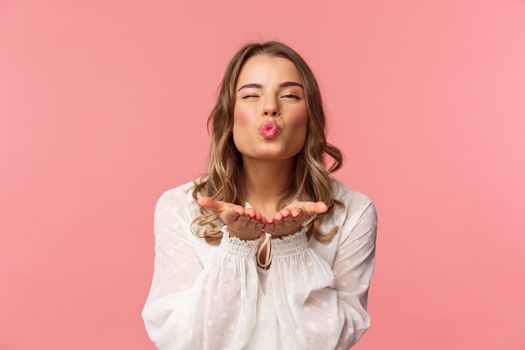 Close-up portrait of lovely feminine girlfriend enjoying spring fine day, sending air kiss at camera, winking and folding lips in mwah, hold hands near mouth, pink background.