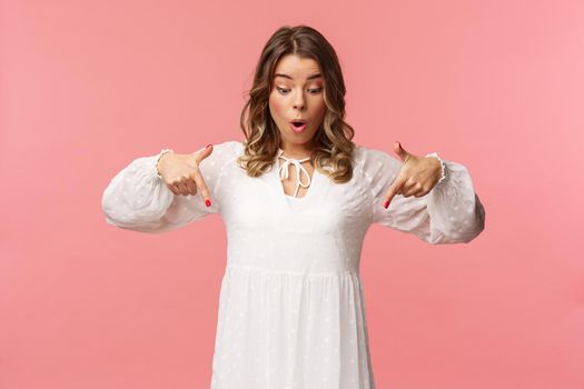 Amazed and wondered good-looking blond european woman 20s, wear beautiful white dress, looking and pointing up, freaking out over something awesome and breathtaking, pink background.