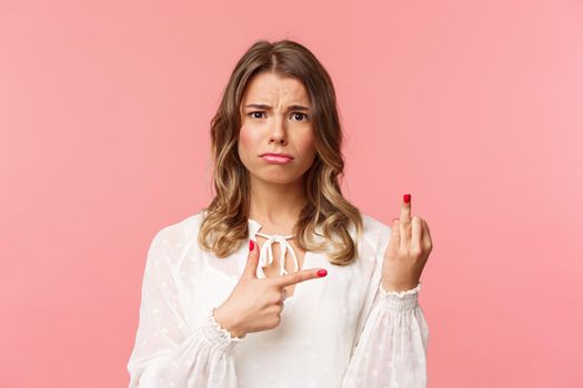 Close-up portrait sad and frustrated young blond girl waited for proposal during romantic date, pointing at finger without weddint ring with puzzled upset expression, standing pink background.