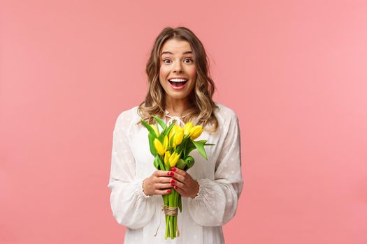 Holidays, beauty and spring concept. Portrait of excited and amazed young beautiful blond girl in white dress, holding yellow tulips, receive flowers as gift, smling amused, pink background.