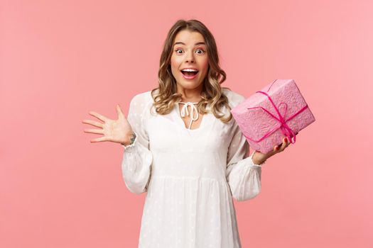 Holidays, celebration and women concept. Portrait of surprised upbeat charismatic blond girl in white dress, spread hands sideways and gasping amazed, receive surprise gift, hold pink box.