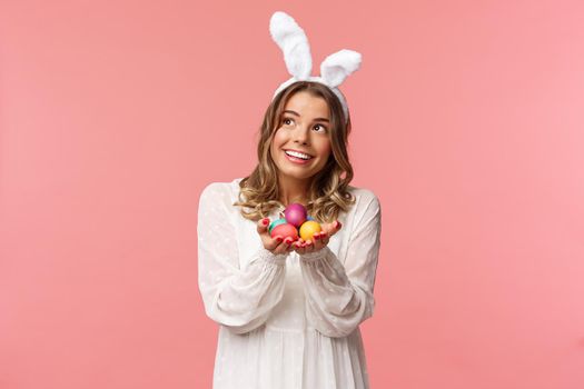 Holidays, spring and party concept. Portrait of cute and tender, lovely blond woman celebrating Easter day look dreamy up with pleased smile, holding easter painted eggs, wear bunny ears.