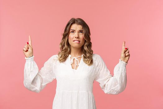 Skeptical and embarrassed blond caucasian woman in white cute dress, grimacing, cringe from seeing something disgusting, frowning doubtful and pointing fingers up strange thing, pink background.
