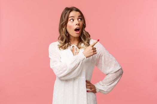 Surprised and excited good-looking blond girl in white cute dress, open mouth in amazement, look curious pointing upper right corner astonished, see something awesome, pink background.