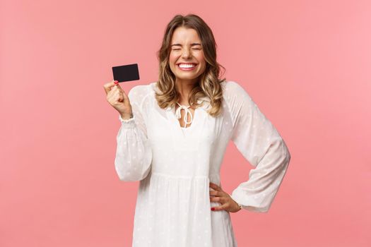 Portrait of excited cute and silly, feminine blond girl in white dress, close eyes giggle and smiling happy, got her first payment new job, holding credit card, use banking service, pink background.