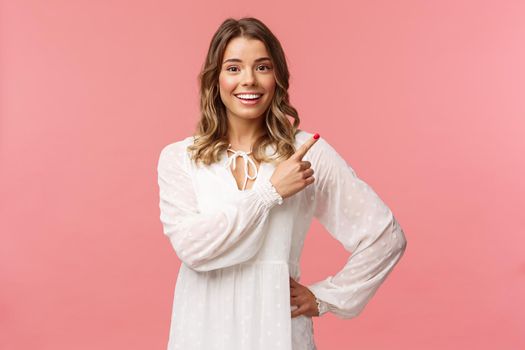 Upbeat good-looking caucasian blond girl in white dress, pointing upper right corner and smiling at camera with interested, curious face discuss special promo spring discounts, pink background.