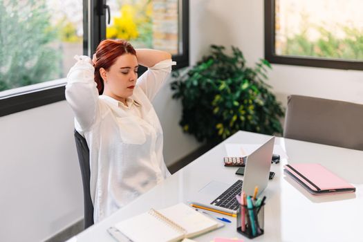 Pretty young woman with red hair massaging her neck while she starts teleworking at home. Self-employed entrepreneur working from home in white shirt. She is working at a light table with a white laptop, calculator, stapler, diaries, notebooks, pens and other devices. Large window in the background with natural light.