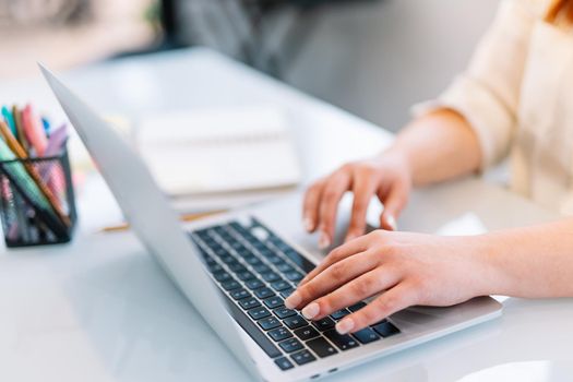 Photo of the hands of a pretty young woman with red hair typing on a laptop while teleworking from home. Young self-employed entrepreneur or businesswoman working from home with white shirt and lots of natural light.