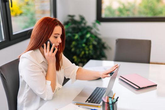 Pretty young beautiful woman with red hair and white shirt teleworking from home talking on her smartphone while using her laptop. She is making a phone call while looking at the laptop screen. Young self-employed businesswoman and entrepreneur working from home with large window in background and natural light.