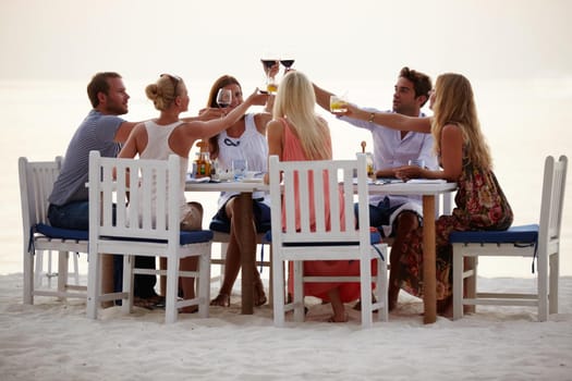 A group of friends toast each other while having dinner on the beach during a summer vacation.