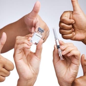 Studio shot of a group of people showing thumbs up and holding the vaccine against a grey background.