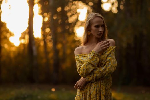 Full body blond female in floral dress looking looking away while standing on grass in evening in forest