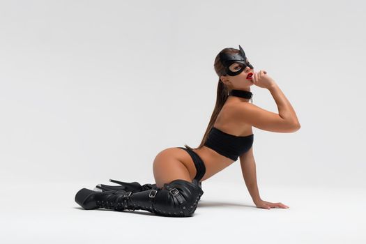 Full body side view of alluring slim tanned brunette in black mask and underwear with high heeled leather boots in white studio