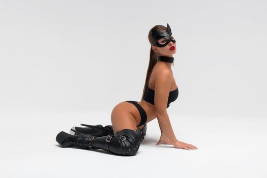 Full body side view of alluring slim tanned brunette in black mask and underwear with high heeled leather boots in white studio