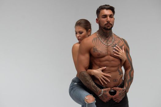 Attractive female embracing handsome shirtless male with tattoos from behind and looking at camera against gray background