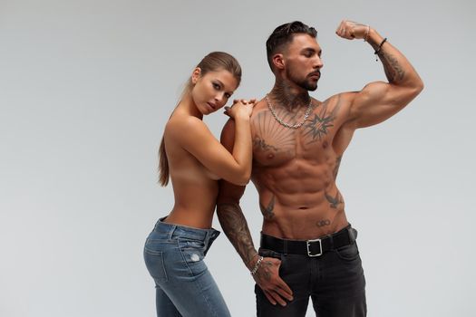 Strong shirtless male embracing and covering breast of attractive topless female on gray background