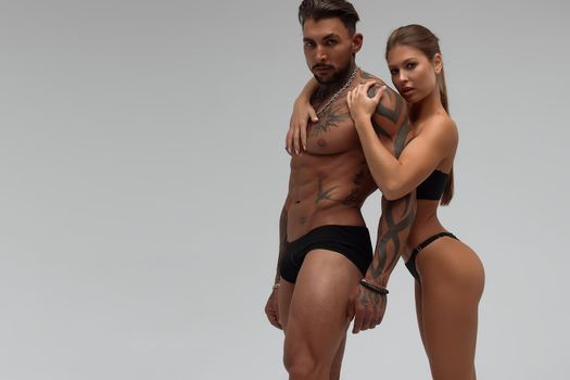 Muscular tattooed man embracing slim topless woman in panties on gray background