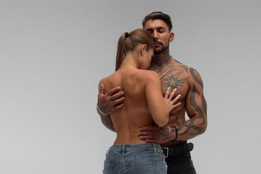 Muscular tattooed man embracing slim topless woman in panties on gray background