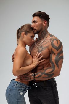 Attractive female embracing handsome shirtless male with tattoos from behind and looking at camera against gray background