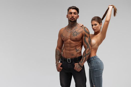 Muscular tattooed man embracing slim topless woman in panties on gray background