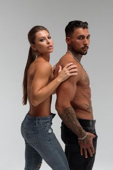 Attractive female embracing handsome shirtless male with tattoos from behind and looking at camera against gray background