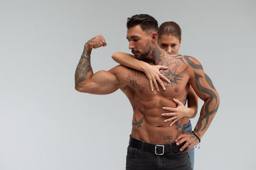 Strong shirtless male embracing and covering breast of attractive topless female on gray background