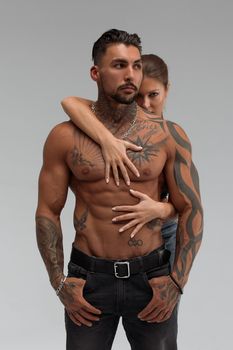 Attractive female embracing handsome shirtless male with tattoos from behind and looking at camera against gray background