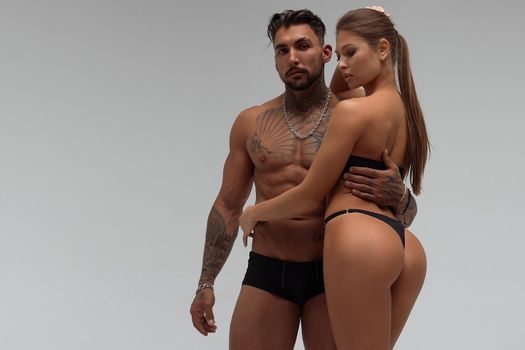 Muscular tattooed man embracing slim topless woman in panties on gray background