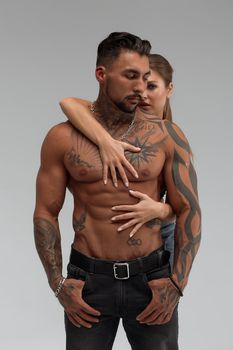 Attractive female embracing handsome shirtless male with tattoos from behind and looking at camera against gray background