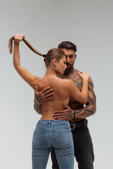 Attractive female embracing handsome shirtless male with tattoos from behind and looking at camera against gray background