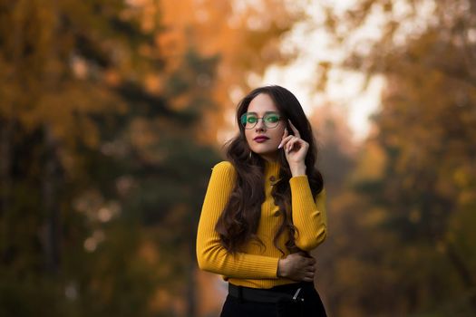 Young long haired brunette in yellow knitted jumper and eyeglasses looking at camera while standing against blurred foliage of autumn trees in park