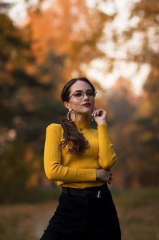 Young long haired brunette in yellow knitted jumper and eyeglasses looking at camera while standing against blurred foliage of autumn trees in park