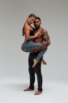 Attractive female embracing handsome shirtless male with tattoos from behind and looking at camera against gray background
