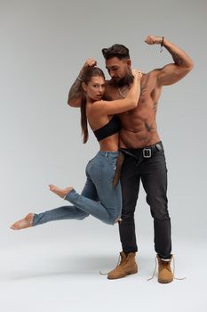 Attractive female embracing handsome shirtless male with tattoos from behind and looking at camera against gray background