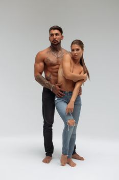 Attractive female embracing handsome shirtless male with tattoos from behind and looking at camera against gray background