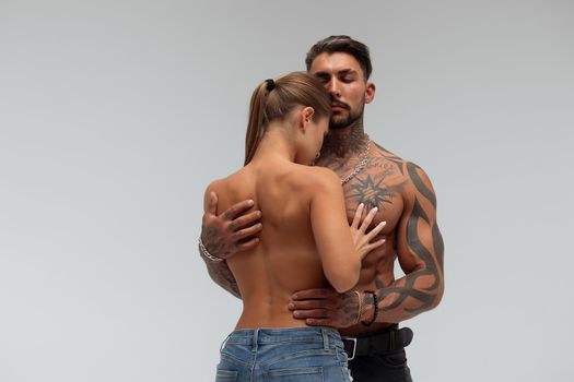 Strong shirtless male embracing and covering breast of attractive topless female on gray background
