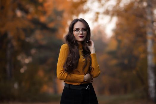 Young long haired brunette in yellow knitted jumper and eyeglasses looking at camera while standing against blurred foliage of autumn trees in park