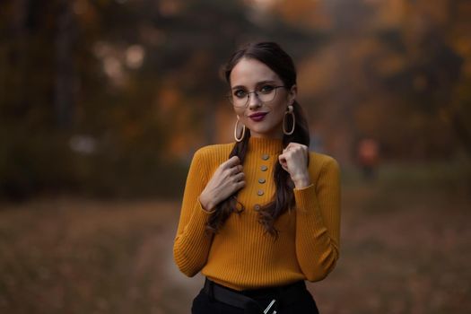 Young long haired brunette in yellow knitted jumper and eyeglasses looking at camera while standing against blurred foliage of autumn trees in park