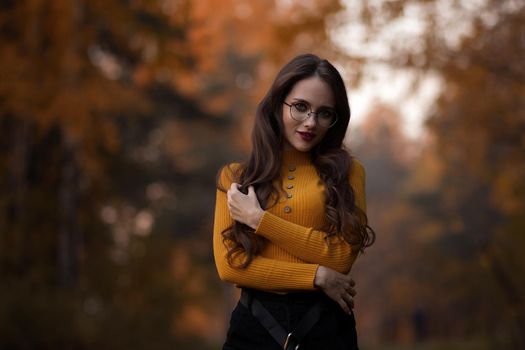 Young long haired brunette in yellow knitted jumper and eyeglasses looking at camera while standing against blurred foliage of autumn trees in park