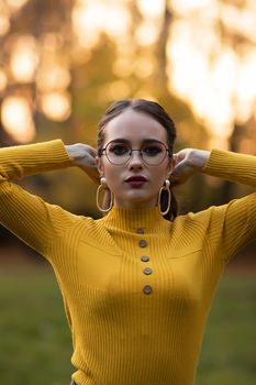 Young long haired brunette in yellow knitted jumper and eyeglasses looking at camera while standing against blurred foliage of autumn trees in park